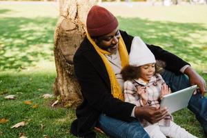 Black grandfather and granddaughter using tablet computer while sitting in park photo