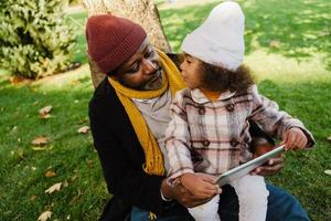 Black grandfather and granddaughter using tablet computer while sitting in park photo