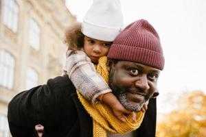 Black girl having fun and sitting on neck of his grandfather in autumn park