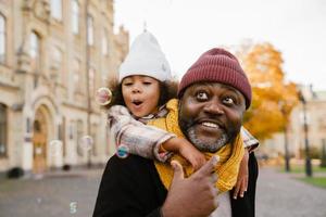 Black girl blowing soap bubbles while walking with grandfather in autumn park photo
