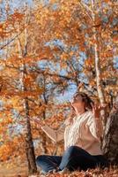 Mujer joven arrojando hojas amarillas en el aire sentado en el bosque de otoño foto