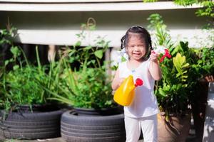 Cute girl was holding yellow watering can, smiling sweetly in garden of small tree in front of house. Children help with housework. Kid play spray nozzle. Summer evenings or spring. 3 year old child.