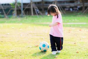 Active kid playing blue-white soccer ball. Girl was happy to play sports to exercise and strengthen her body. Asian children wear white sport shoes. On green grass field. Cute child aged 3-4 years old photo