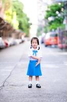 Portrait of Asian pupils girl wearing blue and white school uniform. Child girl get a little embarrassed when your parents take a photo at first baby go to school. Sweet smiling kid aged 3 years old.
