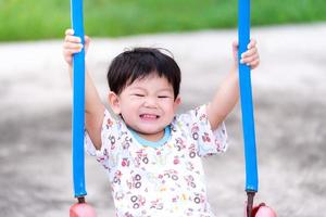 Boy plays on the swing, smiling sweetly. Toddler two hands caught on the blue string of the swings. Little children have fun in the playground. Child is 2 years old. photo