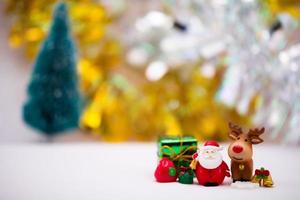 Santa Claus resin dolls stand alongside brown reindeer. The side are decorated with gift boxes, snow flakes and gold bells. Soft white floor. Faint bokeh on gold and silver backdrop and Christmas tree photo