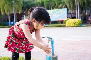 linda niña con mascarilla de tela se está lavando las manos con agua limpia del grifo en el área pública después de hacer su actividad. niño de 4 años. prevenir la propagación de la enfermedad por coronavirus covid 19. foto