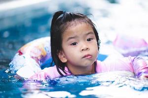 4-5 years old cute Asian girl is swimming using a rubber ring, a child swims around in the water. Children was wondering something in front of her. Kid wear long-sleeved swimsuits. photo