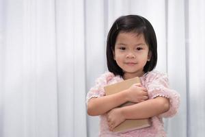 concepto del día mundial del libro y el derecho de autor. una linda niña abraza un libro cerca de su cuerpo. a los niños les encanta leer. adorable niño de 4-5 años. sobre fondo de cortina blanca. educación. De vuelta a la escuela. foto