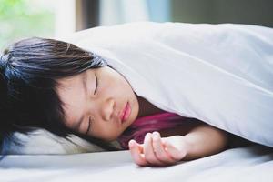 muchacha asiática del niño que duerme en la cama blanda blanca. por la mañana en horario de verano. lindos niños acostados y descansar en paz. niño de 4-5 años. foto