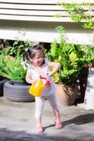 Adorable girl holding yellow watering can peeking at something. Child smiles sweet bright. Happy children help their parents with housework. In evening the sun is warm. Summer or spring. Kids 3-4 year photo