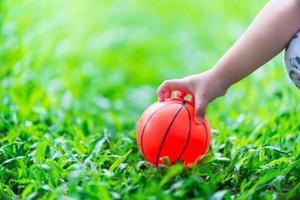 The child's hand is picking up the ball that has fallen on the green grass. Orange ball on the lush bright lawn. photo