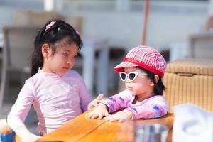 Adorable two Asian girls stand and talk. Child wear sunglasses and a hat. Children go to the sea. Female wear swimsuits. Kid sticks to a table in a restaurant. Baby aged 4-5 years. photo