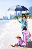 Vertical image. Family travel at the sea and beach. Aunt and granddaughters playing by seaside. Woman holding black umbrella protect hot sun. Summer day. Happy kid girl wear hat and sitting on sand.