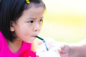 Head shot of Asian kid girl sucking sweet Coconut Smoothie. Mom holding glass to feeding daughter. Baby drinking cool water with tube. Summer time, hot day. Copy space. Children aged 4-5 years old. photo