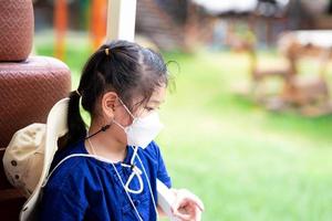 Child wear white face masks 4D shapes to prevent spread of virus and prevent PM2.5 air pollution. Cute kid sits down after playing until her is tired, girl is feeling sad. photo