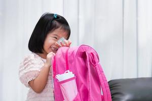 linda niña se ríe mientras empaca sus mochilas escolares para la escuela al comienzo del semestre para que los estudiantes puedan regresar a la escuela como de costumbre después de la epidemia de neumonía por coronavirus. niño sosteniendo una botella de gel de alcohol foto