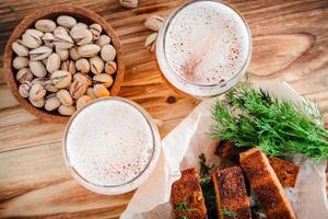 dos vasos de cerveza, tostadas con costra, eneldo y pistachos en una mesa de madera. enfoque selectivo en espuma foto