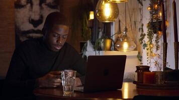 Man sitting at table watching and typing on smartphone and laptop drinking water video