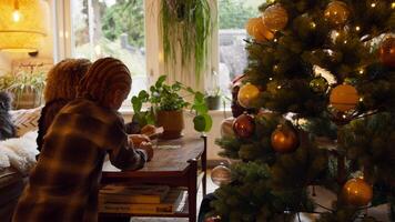 Mujer, niña y hombre con sombrero de navidad jugando a las cartas en la mesa junto al árbol de navidad video