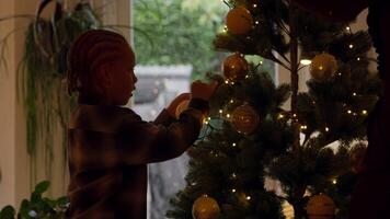 Boy decorating Christmas tree with bauble whilst woman watches video