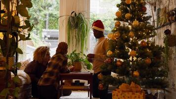 Mujer, niña y hombre con sombrero de navidad jugando a las cartas en la mesa junto al árbol de navidad video