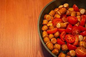 Fried sausages with tomato in process of frying in a frying pan at the red wood table with copy space for text. photo