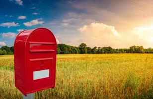 Big red cozy postbox with white empty note space for address is standing outdoor in front of beautiful countryside landscape at sunset background with farm field and poppies flowers. photo