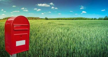 Big red modern postbox with white empty note space for address is standing outdoor in front of beautiful countryside landscape background with farm green wheat field and blue sky. photo