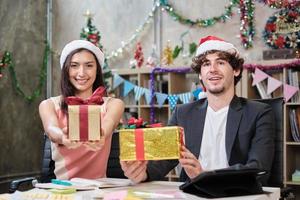 Retrato del equipo de dos compañeros de trabajo mirando a la cámara y dando regalos mientras el negocio trabaja antes de las vacaciones de la empresa en el lugar de trabajo de la oficina, decorado para una celebración del festival de Navidad y una fiesta de año nuevo. foto