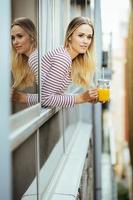 mujer joven bebiendo un vaso de jugo de naranja natural, asomado a la ventana de su casa.