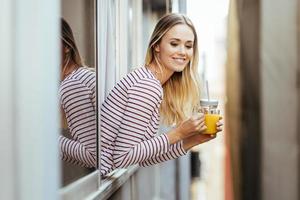 mujer sonriente bebiendo un vaso de jugo de naranja natural, asomado a la ventana de su casa.