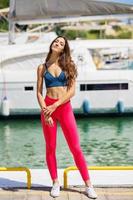 Fitness model in red sportswear outfit posing on waterfront harbour.