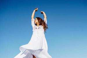 Young woman raising her arms in a beautiful white dress against a blue sky photo