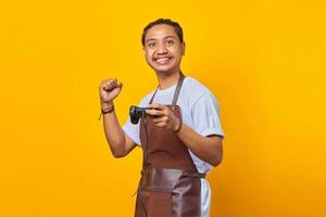 Portrait of handsome asian man wearing apron playing games with joystick make winner gesture isolated on yellow background photo