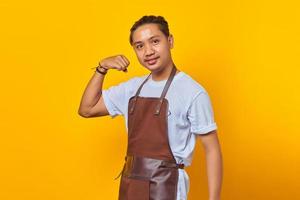 Retrato de sonriente joven asiático vistiendo delantal mostrando los músculos del brazo mientras sonríe con orgullo sobre fondo amarillo foto