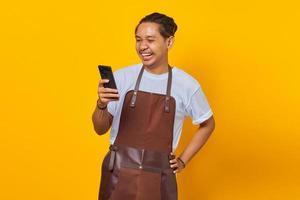 Portrait of laughing handsome Asian young man wearing apron looking at message on smartphone isolated on yellow background photo