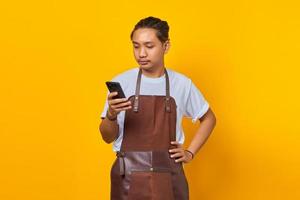 Portrait of handsome asian man wearing apron looking unhappy reading incoming messages on mobile on yellow background photo