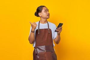 Portrait of smiling handsome man holding cell phone looking sideways with finger pointing at empty space over yellow background photo