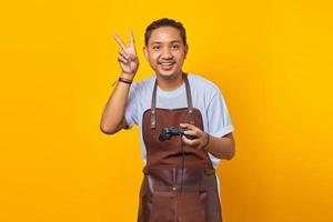 Retrato de alegre apuesto joven asiático vistiendo delantal sosteniendo el controlador del juego y mostrando el signo de la paz sobre fondo amarillo foto