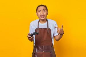 Portrait of surprised handsome Asian young man wearing apron holding game controller and pointing at himself over a yellow background photo