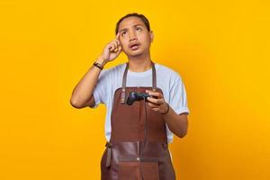Retrato de un apuesto joven asiático vistiendo delantal sosteniendo el controlador del juego y pensando en algo aislado sobre fondo amarillo foto