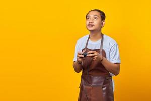 Portrait of asian man wearing apron with tense expression playing game with joystick isolated on yellow background photo