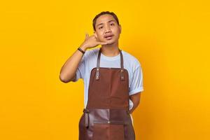 Portrait of Asian young man wearing apron making call sign with dislike expression on yellow background photo