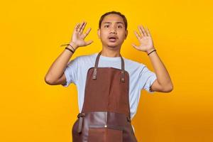 Shocked and surprised asian young man wearing an apron looks forward with his mouth open and both hands raised photo