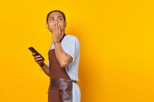 Shocked asian man covering mouth with hands and holding smartphone over yellow background photo