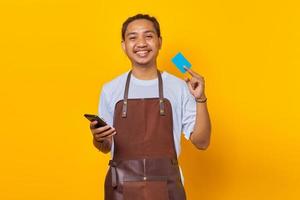 Portrait of smiling handsome barista showing credit card to pay and holding smartphone photo