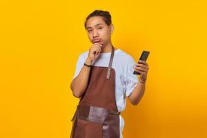 Handsome Asian young man wearing apron holding cell phone and pensive thinking about something with hand on chin photo