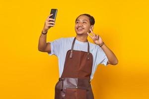 Portrait of asian man smiling and showing peace sign while taking selfie on mobile isolated on yellow background photo