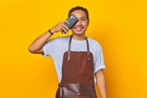 Portrait of smiling handsome Asian young man wearing apron closing eyes with smartphone isolated on yellow background